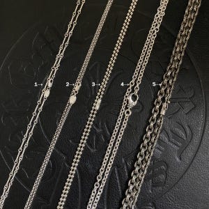 May include: Five silver necklaces with different chain styles are displayed against a dark background. The chains vary in design, including delicate link chains, ball chains, and thicker, textured chains. Each necklace has a clasp or connector.
