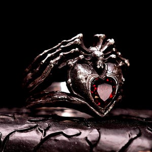 May include: A gothic-style ring with a heart-shaped red gemstone. The ring is made of dark metal and features intricate skeletal and spiderweb-like details. The heart-shaped stone is a deep red color, set in a detailed metal frame.
