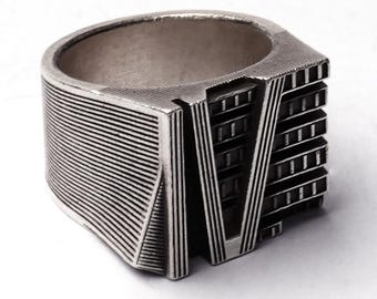 May include: A silver-colored ring with a unique architectural design. The ring features a rectangular shape with horizontal and vertical lines, resembling a building facade. The ring's design includes a V-shaped element and a series of rectangular cutouts.