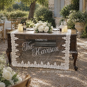 May include: A rectangular white lace table runner with scalloped edges draped over a dark wooden table. The runner features the white printed text "CHEERS TO The Harrison" and measures 50 inches by 70 inches, or 127 cm by 178 cm.