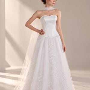 May include: A white strapless wedding dress featuring a structured satin bodice with a sweetheart neckline and a full-length skirt layered with sheer tulle and intricate floral lace appliqués, accompanied by a matching sheer tulle wrap.