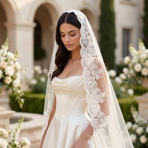 May include: A white wedding veil featuring intricate floral lace trim along the edges, draped over a satin corset-style gown with a sweetheart neckline. The sheer tulle veil extends downward, showcasing detailed rose-patterned embroidery.