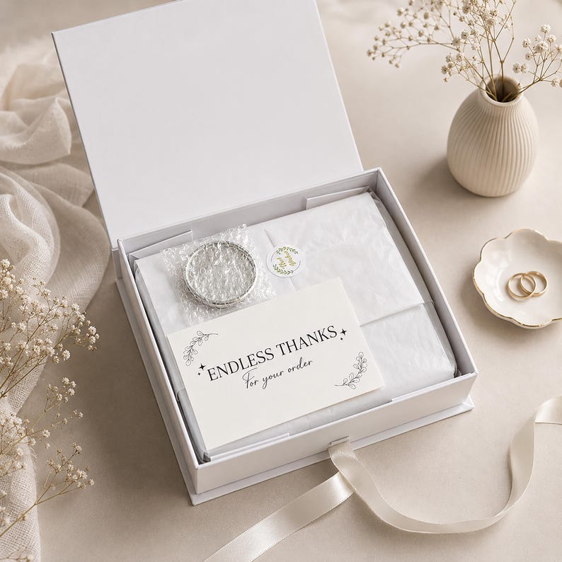 Puede incluir: Una caja de regalo rectangular blanca con tapa abatible, que contiene papel de seda blanco, un art&iacute;culo circular de tono plateado envuelto en pl&aacute;stico de burbujas y una tarjeta blanca impresa con el texto ENDLESS THANKS For your order.