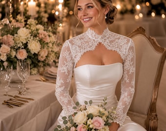 May include: A white strapless gown featuring a high-neck lace overlay with long sleeves. The bodice is made of smooth satin, while the lace overlay displays intricate floral patterns. The garment is worn with a bouquet of white and pink roses.