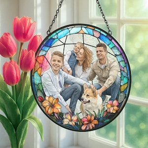 May include: A round, stained-glass window hanging with a family portrait. The window has a black metal frame and a chain for hanging. The family includes two parents, two children, and a dog. The window is surrounded by colorful flowers.