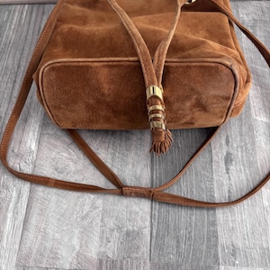 Puede incluir: Un bolso tipo cubo de ante marr&oacute;n con una correa larga y ajustable. El bolso presenta detalles met&aacute;licos dorados en la parte superior y en el cierre de cord&oacute;n. El bolso tiene una base rectangular y un aspecto suave y texturizado.