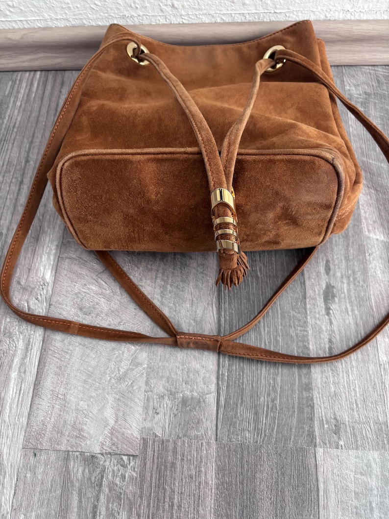 Puede incluir: Un bolso tipo cubo de ante marr&oacute;n con una correa larga y ajustable. El bolso presenta detalles met&aacute;licos dorados en la parte superior y en el cierre de cord&oacute;n. El bolso tiene una base rectangular y un aspecto suave y texturizado.