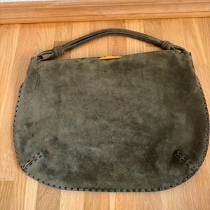 May include: Olive green suede handbag with a rounded shape and a top handle. The bag features a contrasting yellow accent and decorative stitching around the edges. The handle is knotted at the top.