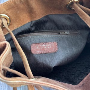 Puede incluir: Un bolso de ante marr&oacute;n con cord&oacute;n y detalles dorados. El interior tiene un forro marr&oacute;n y un bolsillo con cremallera. Una etiqueta de cuero en el interior dice "Valentino Garavani".