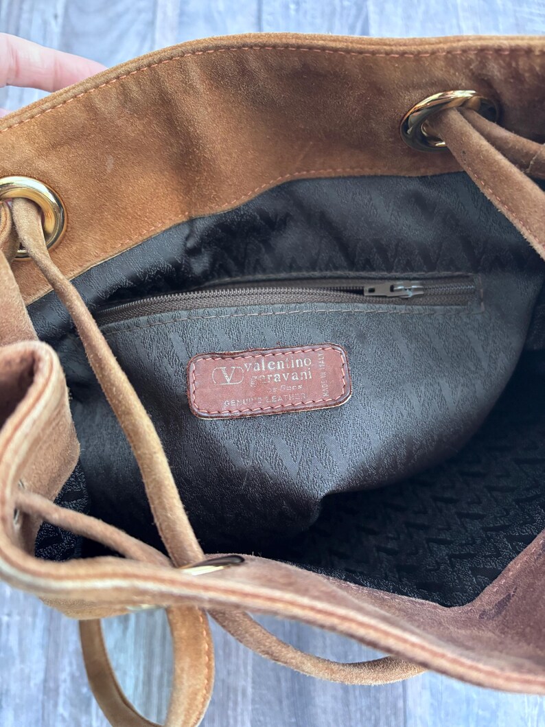Puede incluir: Un bolso de ante marr&oacute;n con cord&oacute;n y detalles dorados. El interior tiene un forro marr&oacute;n y un bolsillo con cremallera. Una etiqueta de cuero en el interior dice "Valentino Garavani".
