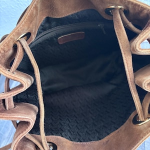 Puede incluir: Un bolso tipo cubo de ante marr&oacute;n con cierre de cord&oacute;n. El interior est&aacute; forrado con una tela oscura y tiene un bolsillo con cremallera. Los ojales y los extremos del cord&oacute;n de metal dorado a&ntilde;aden detalles al dise&ntilde;o.