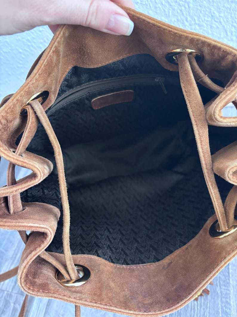 Puede incluir: Un bolso tipo cubo de ante marr&oacute;n con cierre de cord&oacute;n. El interior est&aacute; forrado con una tela oscura y tiene un bolsillo con cremallera. Los ojales y los extremos del cord&oacute;n de metal dorado a&ntilde;aden detalles al dise&ntilde;o.