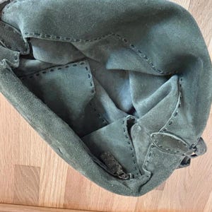May include: Olive green suede handbag with visible stitching details. The bag features a zipper closure and a flap with a buckle. The interior is visible, showing the soft suede material. The bag is on a wooden surface.