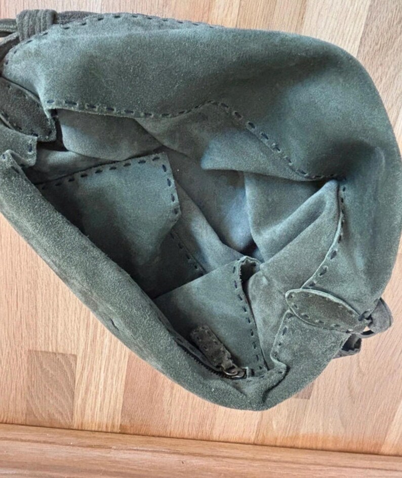 May include: Olive green suede handbag with visible stitching details. The bag features a zipper closure and a flap with a buckle. The interior is visible, showing the soft suede material. The bag is on a wooden surface.