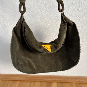 May include: Olive green suede shoulder bag with a curved design. The bag features a yellow triangle logo and a stitched trim. The strap is made of the same material as the bag. The bag is hanging against a white wall.