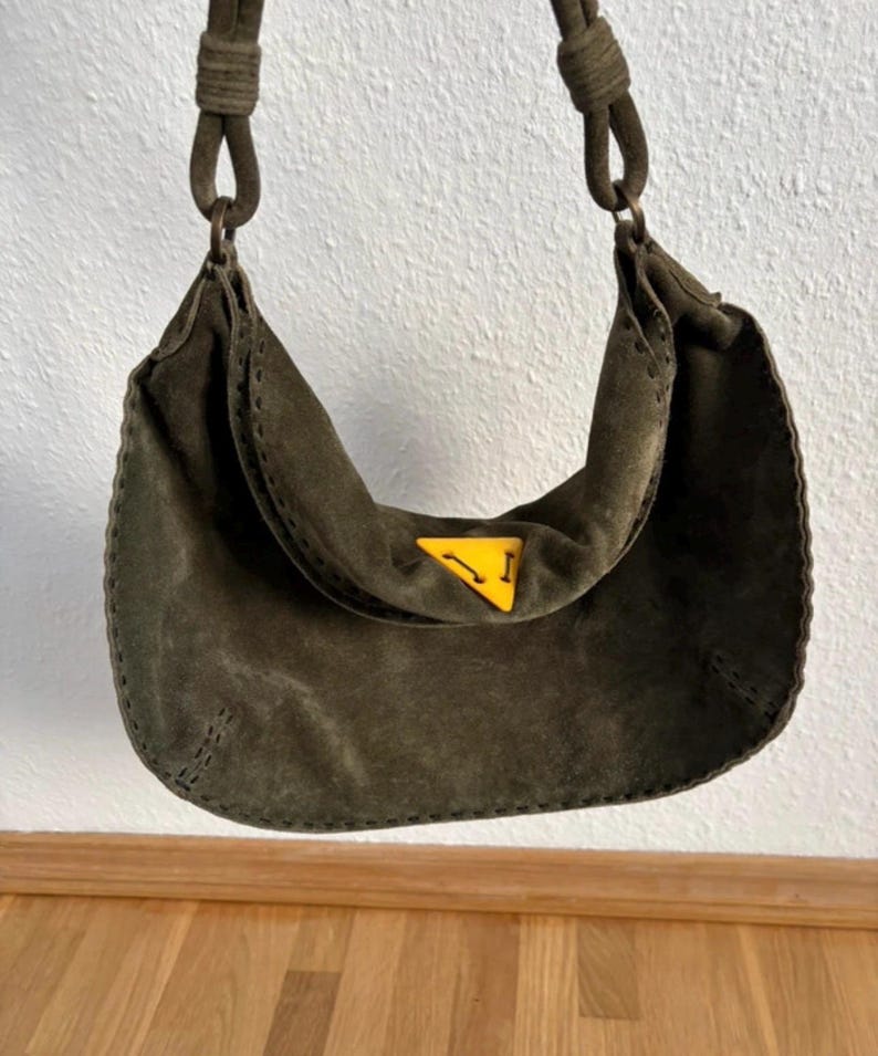 May include: Olive green suede shoulder bag with a curved design. The bag features a yellow triangle logo and a stitched trim. The strap is made of the same material as the bag. The bag is hanging against a white wall.