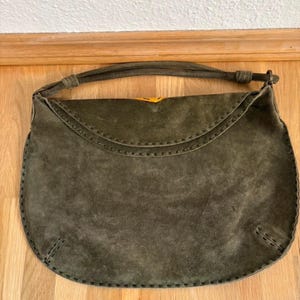 May include: Olive green suede handbag with a curved flap and stitched detailing. The bag has a matching handle and a yellow clasp. The bag is sitting on a wooden surface.