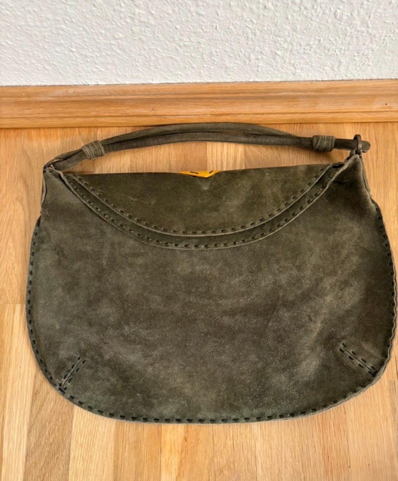 May include: Olive green suede handbag with a curved flap and stitched detailing. The bag has a matching handle and a yellow clasp. The bag is sitting on a wooden surface.