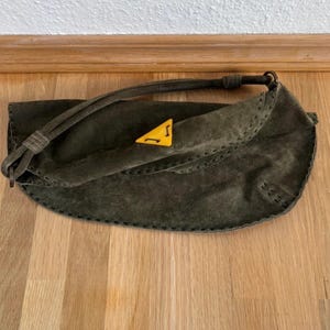 May include: Olive green suede handbag with a curved shape and a long strap. The bag features a yellow triangle emblem with a black design. The edges are stitched with dark thread. The bag is on a wooden surface.
