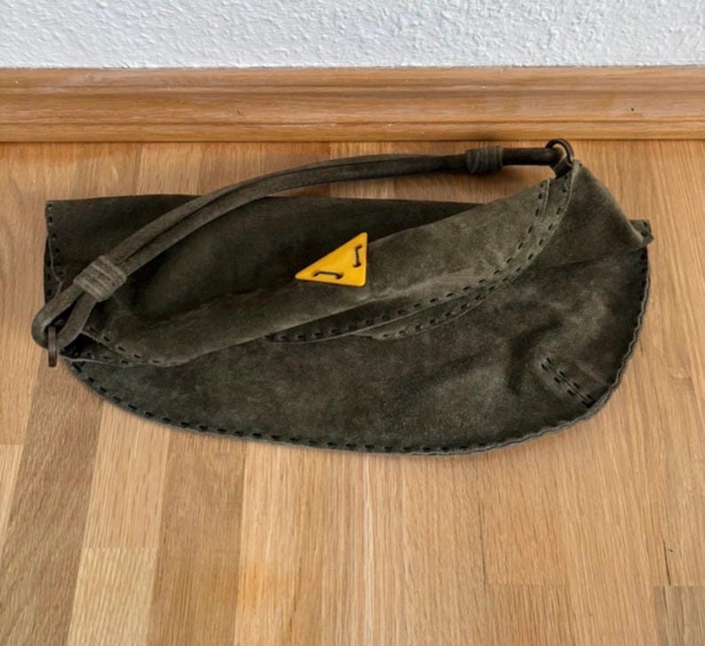 May include: Olive green suede handbag with a curved shape and a long strap. The bag features a yellow triangle emblem with a black design. The edges are stitched with dark thread. The bag is on a wooden surface.