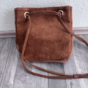 Puede incluir: Bolso de hombro de ante marr&oacute;n con cierre de cord&oacute;n y una correa larga y fina. El bolso tiene forma rectangular con esquinas redondeadas y ojales met&aacute;licos dorados. El bolso est&aacute; hecho de un material suave y texturizado.