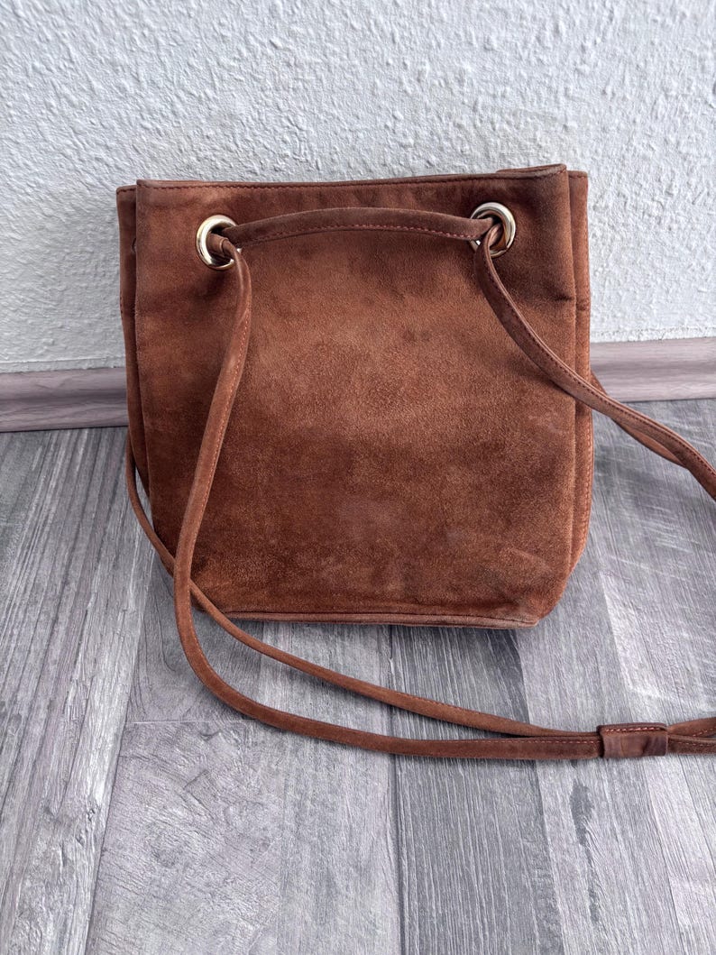 Puede incluir: Bolso de hombro de ante marr&oacute;n con cierre de cord&oacute;n y una correa larga y fina. El bolso tiene forma rectangular con esquinas redondeadas y ojales met&aacute;licos dorados. El bolso est&aacute; hecho de un material suave y texturizado.