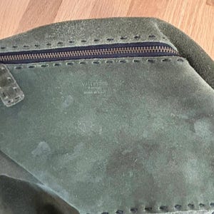 May include: A green suede handbag with a zipper closure and stitched detailing. The bag features the brand name "VALENTINO" and "MADE IN ITALY" printed on the interior. The bag is a muted green color.