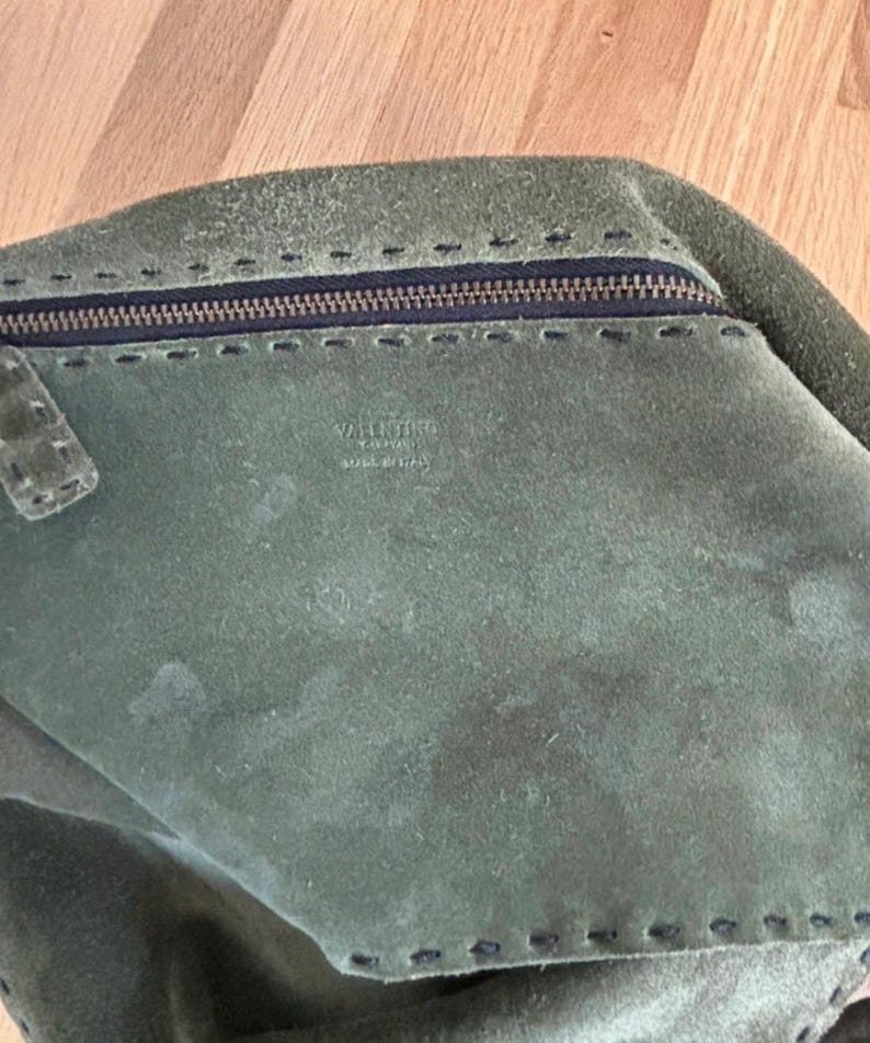 May include: A green suede handbag with a zipper closure and stitched detailing. The bag features the brand name "VALENTINO" and "MADE IN ITALY" printed on the interior. The bag is a muted green color.