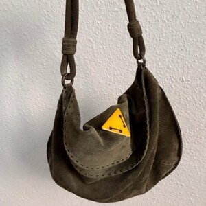 May include: Olive green suede shoulder bag with a rounded shape and a knotted strap. The bag features a yellow triangle accent with black stitching. The bag is held up by a dark brown strap.