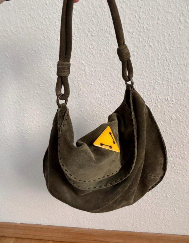 May include: Olive green suede shoulder bag with a rounded shape and a knotted strap. The bag features a yellow triangle accent with black stitching. The bag is held up by a dark brown strap.