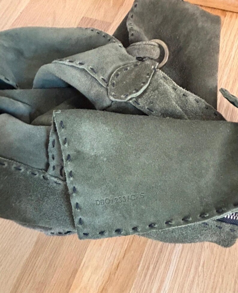 May include: Close-up of a sage green suede bag with visible stitching and a metal ring detail. The bag features a flap closure and a textured surface. The text "DBO/233/C/S" is stamped on the suede.