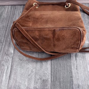 Puede incluir: Un bolso tipo cubo de ante marr&oacute;n con cierre de cord&oacute;n y correas para el hombro. El bolso tiene un fondo redondeado y ojales met&aacute;licos dorados. El bolso est&aacute; hecho de un material suave y texturizado.