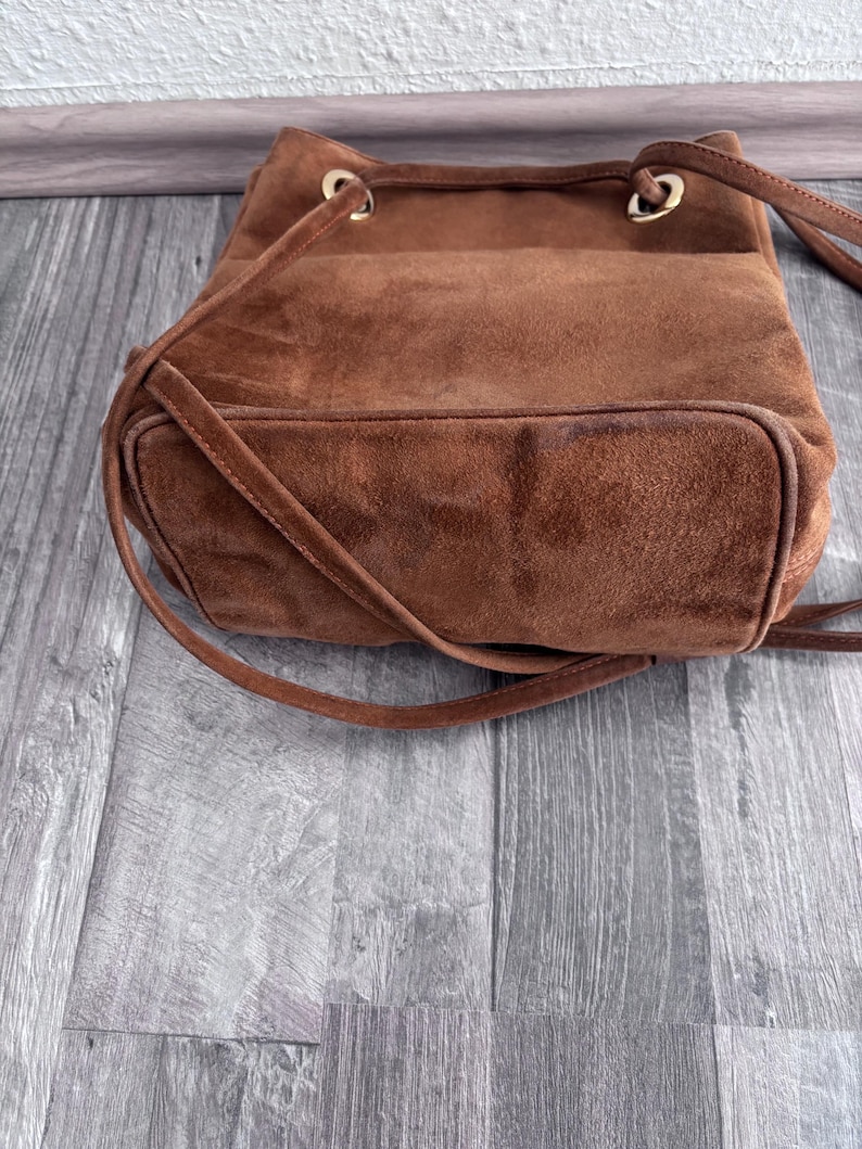 Puede incluir: Un bolso tipo cubo de ante marr&oacute;n con cierre de cord&oacute;n y correas para el hombro. El bolso tiene un fondo redondeado y ojales met&aacute;licos dorados. El bolso est&aacute; hecho de un material suave y texturizado.