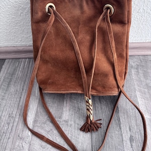 Puede incluir: Bolso de hombro de ante marr&oacute;n con forma rectangular. El bolso presenta ojales met&aacute;licos dorados y una borla decorativa con un adorno met&aacute;lico dorado. El bolso tiene correas largas y finas.