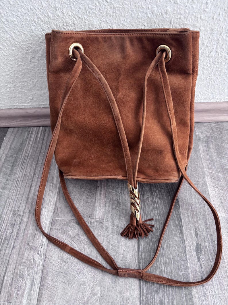 Puede incluir: Bolso de hombro de ante marr&oacute;n con forma rectangular. El bolso presenta ojales met&aacute;licos dorados y una borla decorativa con un adorno met&aacute;lico dorado. El bolso tiene correas largas y finas.