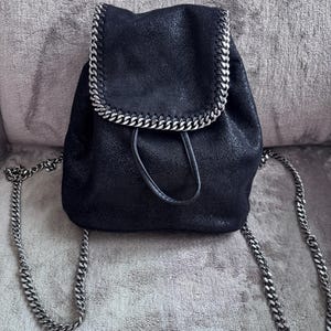 May include: Black backpack with a silver chain trim and straps. The bag has a drawstring closure and a flap. The material appears to be textured. The backpack is sitting on a gray surface.