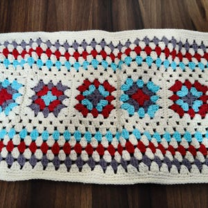 Peut inclure: Un chemin de table rectangulaire en crochet avec un motif répétitif de carrés granny. Le design intègre des fils de couleur crème, rouge vif, bleu clair et gris foncé disposés en un motif géométrique avec une bordure festonnée.