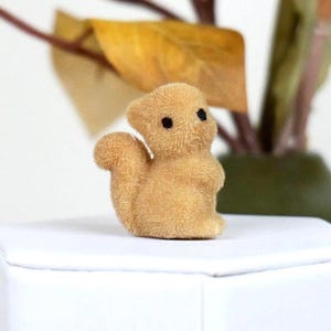 May include: A small, tan-colored squirrel figurine with a fluffy texture. The squirrel has black eyes and a curved tail. It is standing on a white surface, with blurred foliage in the background.