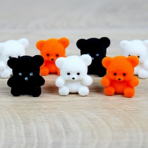 May include: A collection of small, fuzzy teddy bears in various colors, including white, black, and orange. The bears have small, dark eyes and are arranged on a light-colored wooden surface. The bears are a cute and whimsical decorative item.
