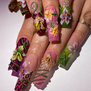 May include: A hand displaying long, sculpted fingernails with elaborate 3D floral designs. The nails feature pink, green, and yellow flowers, leopard print accents, and swirling green vines. A gold ring with a pearl is visible.