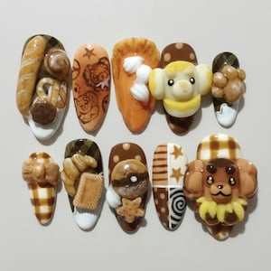 May include: A set of ten artificial nails with detailed, 3D designs. The nails feature various food-themed decorations, including bread, cookies, and a dog. Colors include brown, white, yellow, and orange.