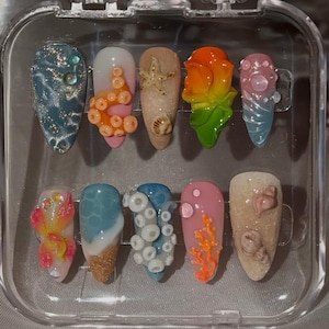 May include: A set of ten artificial nails with various ocean-themed designs. The nails feature colors like blue, pink, and beige, with embellishments such as starfish, coral, and octopus tentacles. The nails are displayed in a clear plastic case.