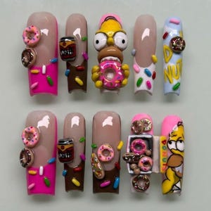 May include: A set of ten long, artificial nails with a variety of designs. Some nails feature donuts, sprinkles, and a cartoon character. Colors include pink, brown, beige, and blue.