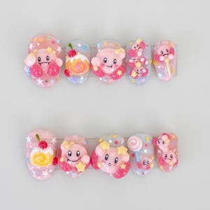 May include: A set of ten artificial nails with a cartoon theme. Each nail features a different design, including pink characters, stars, and sweets. The nails are primarily pink, with accents of yellow, red, and blue.