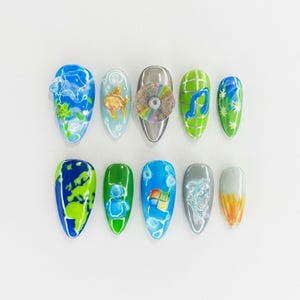 Könnte beinhalten: Zehn künstliche Fingernägel mit verschiedenen Designs. Die Designs umfassen eine CD, Noten, einen Goldfisch, ein Windows-Logo und abstrakte Kunst. Die Nägel sind in Blau, Grün, Silber und Orange bemalt.