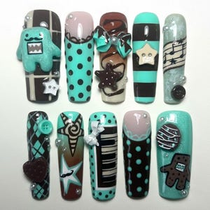 May include: A collection of ten artificial nails with various designs. The nails feature a mix of colors including teal, brown, and pink, with decorative elements like polka dots, bows, stars, and musical notes. Some nails have 3D embellishments.