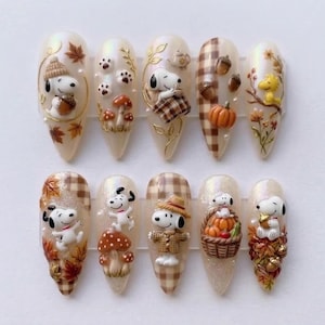 May include: A set of ten press-on nails featuring autumn-themed 3D designs. The nails display Snoopy and Woodstock characters, pumpkins, mushrooms, acorns, and fall leaves in shades of brown, orange, and cream with plaid and glitter accents.