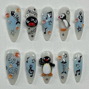May include: A set of ten long, oval-shaped acrylic nails with various designs. The nails feature a light blue base with fish bones, musical notes, stars, and penguin-themed art. Some nails have the word "NOOT" and orange embellishments.