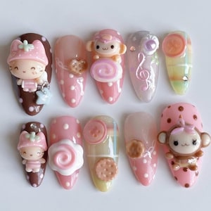 May include: A set of ten press-on nails featuring 3D charms including cartoon characters, pink and brown polka dots, buttons, cookies, and a musical treble clef. The designs incorporate pastel colors, glossy finishes, and small decorative rhinestones.