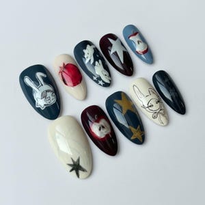 K&ouml;nnte beinhalten: Ein Set aus zehn k&uuml;nstlichen N&auml;geln mit verschiedenen Designs. Die N&auml;gel zeigen Illustrationen eines Kaninchens, Sterne und &Auml;pfel in Farben wie Marine, Wei&szlig;, Rot und Gold. Die N&auml;gel sind oval geformt und auf einer wei&szlig;en Oberfl&auml;che angeordnet.